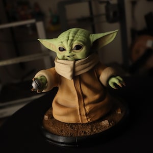 Puede incluir: Una figura de criatura de piel verde con grandes orejas puntiagudas y ojos oscuros, que viste una t&uacute;nica color canela con cuello alto. La figura sostiene un peque&ntilde;o objeto esf&eacute;rico plateado en una mano y se apoya en una base circular.