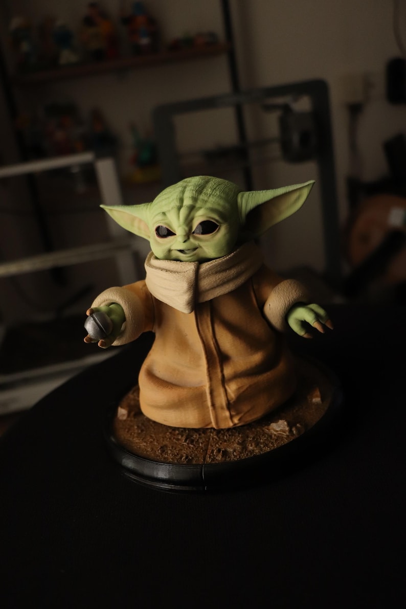 Puede incluir: Una figura de criatura de piel verde con grandes orejas puntiagudas y ojos oscuros, que viste una t&uacute;nica color canela con cuello alto. La figura sostiene un peque&ntilde;o objeto esf&eacute;rico plateado en una mano y se apoya en una base circular.