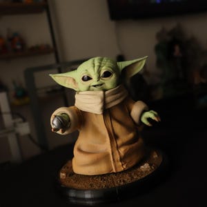 Puede incluir: Una figura de criatura de piel verde con grandes orejas puntiagudas y ojos oscuros, que viste una t&uacute;nica color canela con cuello alto. La figura sostiene un peque&ntilde;o objeto esf&eacute;rico plateado en una mano y se apoya sobre una base marr&oacute;n.