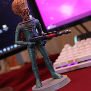 Puede incluir: Una figura de un marciano de Mars Attacks con una cabeza grande, texturizada y similar a un cerebro, ojos muy abiertos y rostro esquelético. El personaje viste un traje espacial de color verde azulado metálico y sostiene una pistola de rayos.
