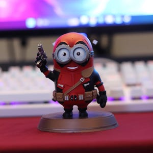 Può includere: Una piccola statuetta di un personaggio che combina un minion giallo con un costume da supereroe rosso e nero. La figura presenta grandi occhiali rotondi, una cintura multiuso con tasche e una piccola pistola giocattolo su una base.
