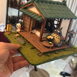 May include: Miniature Japanese-style house diorama featuring characters from the animated film "My Neighbor Totoro." The scene includes Totoro, No-Face, and a Kodama figure, with a green roof and wooden porch. The base is covered in artificial grass.