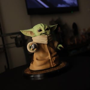 Puede incluir: Una figura de criatura verde con orejas grandes y ojos oscuros, que lleva una t&uacute;nica color canela con cuello alto. La figura est&aacute; sobre una base circular marr&oacute;n y sostiene un peque&ntilde;o objeto esf&eacute;rico plateado en su mano derecha, extendida.