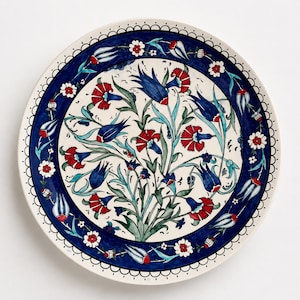 Op de afbeelding: Een ronde, decoratieve keramische plaat met een witte achtergrond en een blauwe rand. De plaat heeft een bloemenontwerp met rode en blauwe bloemen en groene bladeren. De rand heeft een geschulpte rand.