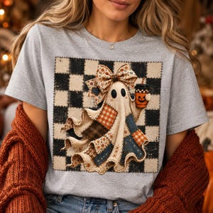 May include: A heather gray t-shirt featuring a graphic of a ghost wearing a large patterned bow and holding an orange pumpkin-themed coffee cup. The ghost is layered over a black and white checkered square background with a stitched border design.