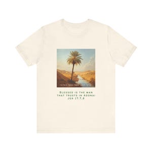 Könnte beinhalten: Cremefarbenes T-Shirt mit einem quadratischen Bild einer Wüstenlandschaft mit einer Palme und einem Fluss. Der Text "BLESSED IS THE MAN THAT TRUSTS IN ADONAI JER 17:7,8" ist darunter gedruckt. Das Shirt besteht aus weichem Stoff.
