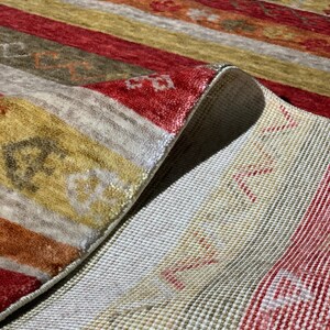 May include: Close-up of a colorful rug with a striped pattern. The rug features alternating bands of red, gold, and beige. The rug's underside is visible, revealing a white woven backing. The rug is folded over, showcasing its texture and design.
