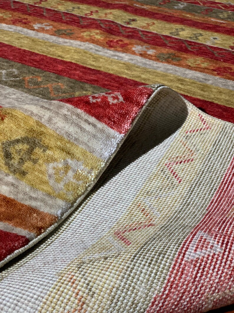 May include: Close-up of a colorful rug with a striped pattern. The rug features alternating bands of red, gold, and beige. The rug's underside is visible, revealing a white woven backing. The rug is folded over, showcasing its texture and design.