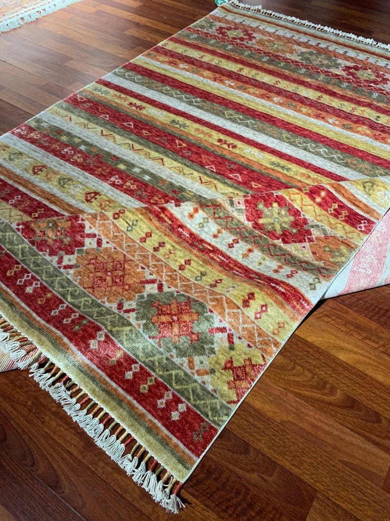 May include: A rectangular area rug with a striped pattern in shades of red, yellow, orange, and green. The rug features a geometric design and fringe along the edges. The rug is placed on a wooden floor.