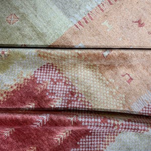 May include: Close-up of a folded textile with a geometric pattern. The fabric features a color palette of beige, peach, and red. The design includes diamond and arrow-like shapes, creating a textured appearance. The textile appears to be soft and possibly woven.