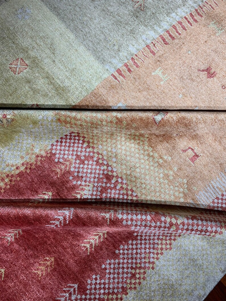 May include: Close-up of a folded textile with a geometric pattern. The fabric features a color palette of beige, peach, and red. The design includes diamond and arrow-like shapes, creating a textured appearance. The textile appears to be soft and possibly woven.