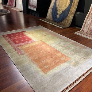 May include: A rectangular area rug with a light gray background and a series of rectangular and square shapes in shades of red, orange, and yellow. The rug has a fringe along the bottom edge and is displayed on a dark wood floor.