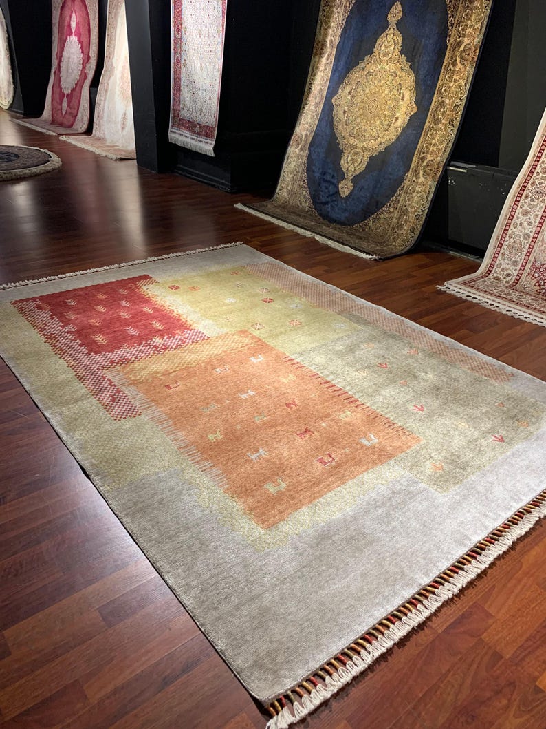 May include: A rectangular area rug with a light gray background and a series of rectangular and square shapes in shades of red, orange, and yellow. The rug has a fringe along the bottom edge and is displayed on a dark wood floor.