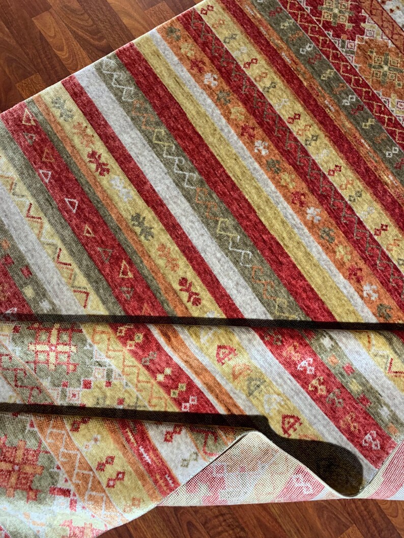 May include: A colorful area rug with a striped pattern in shades of red, yellow, green, and gray. The rug features geometric designs and is partially rolled up, revealing the underside.
