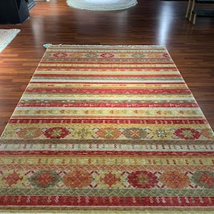May include: A rectangular rug with a striped pattern in shades of red, yellow, and green. The rug features geometric designs and a fringed edge. The rug is displayed on a wooden floor.