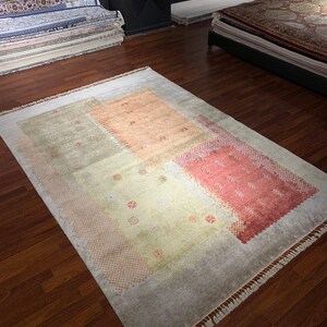 May include: A rectangular area rug with a light gray border and a central design of muted peach, green, and coral blocks. The rug has a fringed edge and is displayed on a hardwood floor. The rug is a decorative floor covering.