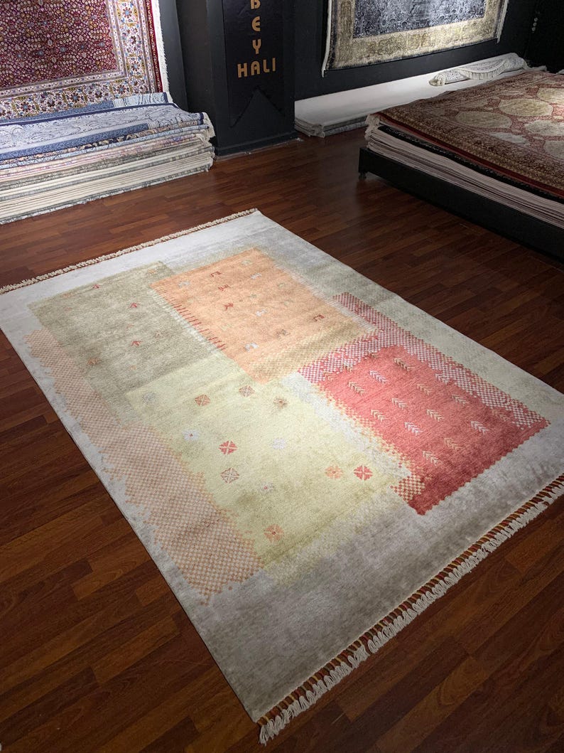 May include: A rectangular area rug with a light gray border and a central design of muted peach, green, and coral blocks. The rug has a fringed edge and is displayed on a hardwood floor. The rug is a decorative floor covering.