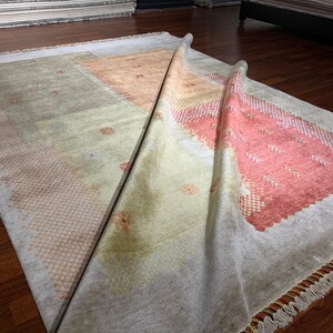 May include: A rectangular rug with a light gray background and geometric patterns in peach and coral. The rug is partially rolled up, revealing a fringed edge and a glimpse of other rugs in the background.