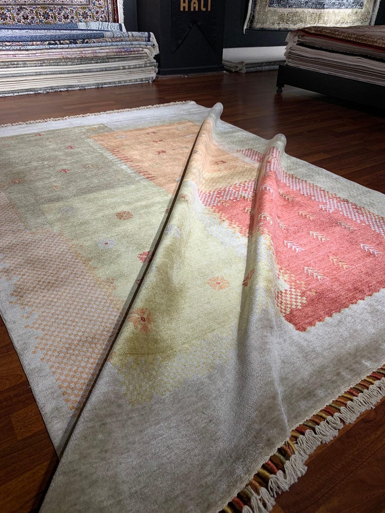 May include: A rectangular rug with a light gray background and geometric patterns in peach and coral. The rug is partially rolled up, revealing a fringed edge and a glimpse of other rugs in the background.