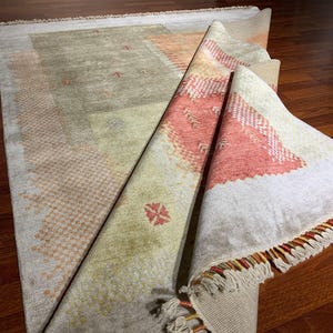 May include: A partially rolled-up area rug with a beige and peach color scheme. The rug features geometric patterns and a fringed edge. The rug is displayed on a hardwood floor, showcasing its texture and design.