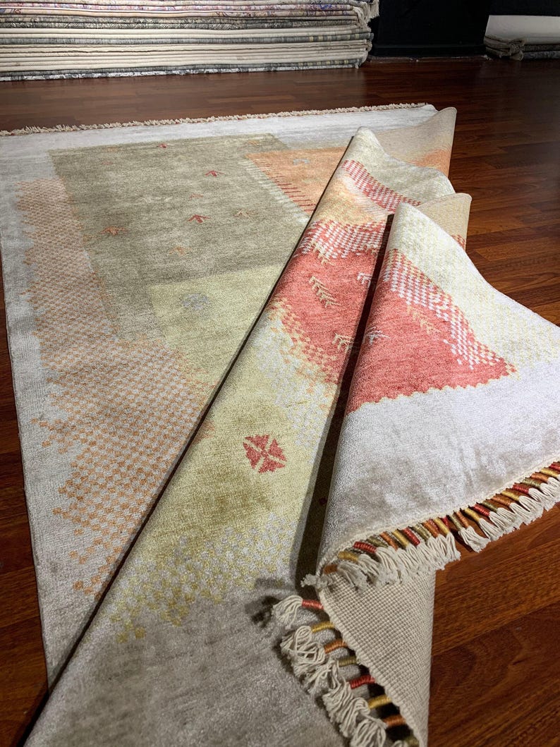 May include: A partially rolled-up area rug with a beige and peach color scheme. The rug features geometric patterns and a fringed edge. The rug is displayed on a hardwood floor, showcasing its texture and design.