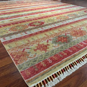 May include: A rectangular area rug with a striped pattern in shades of red, yellow, and green. The rug features a fringe along one edge and intricate geometric designs within the stripes. The rug is placed on a wooden floor.