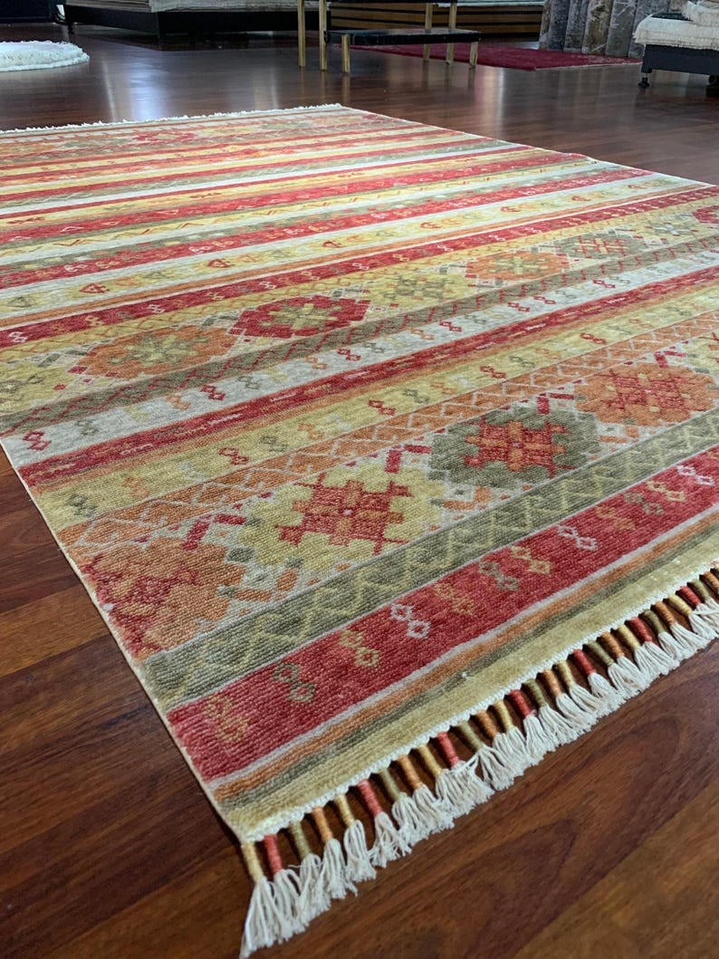 May include: A rectangular area rug with a striped pattern in shades of red, yellow, and green. The rug features a fringe along one edge and intricate geometric designs within the stripes. The rug is placed on a wooden floor.