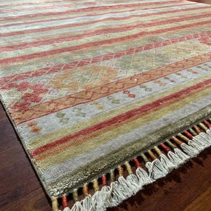 May include: A rectangular rug with a striped pattern in shades of red, yellow, and green. The rug has a fringe along one edge and a detailed geometric design in the center. The rug is placed on a dark wooden floor.