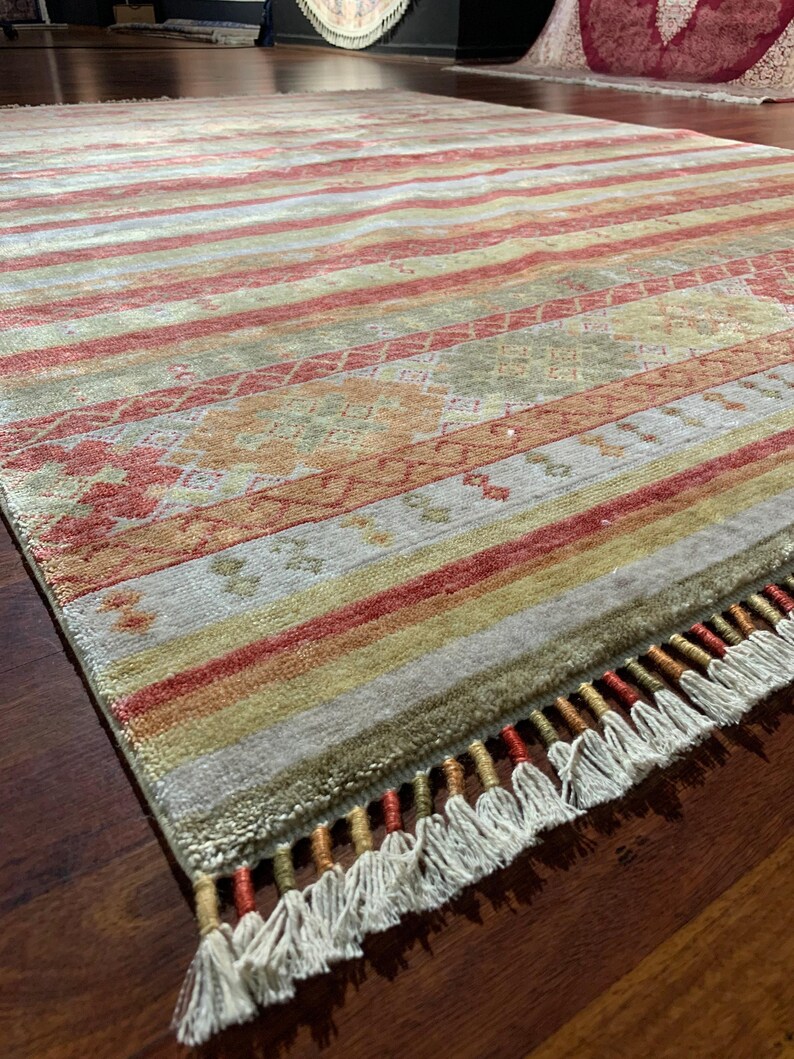 May include: A rectangular rug with a striped pattern in shades of red, yellow, and green. The rug has a fringe along one edge and a detailed geometric design in the center. The rug is placed on a dark wooden floor.