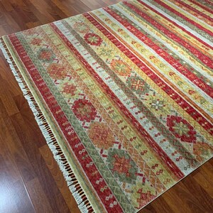 May include: A rectangular area rug with a striped pattern in shades of red, yellow, green, and beige. The rug features geometric designs and fringe on the edges. The rug is placed on a hardwood floor.