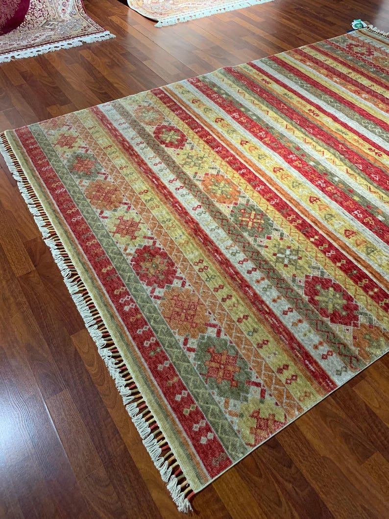 May include: A rectangular area rug with a striped pattern in shades of red, yellow, green, and beige. The rug features geometric designs and fringe on the edges. The rug is placed on a hardwood floor.