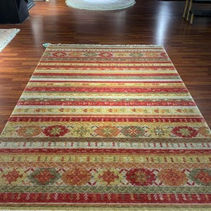 May include: A rectangular rug with a striped pattern in shades of red, yellow, and green. The rug features geometric designs and a fringed edge. The rug is displayed on a wooden floor.