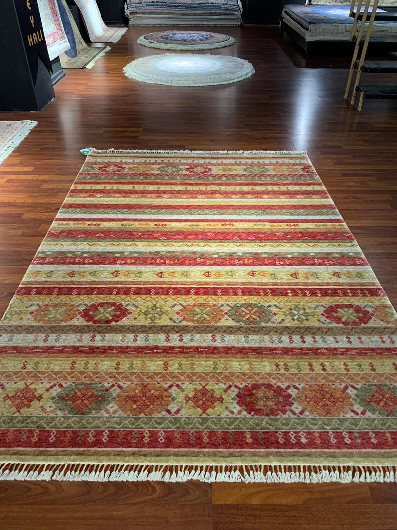 May include: A rectangular rug with a striped pattern in shades of red, yellow, and green. The rug features geometric designs and a fringed edge. The rug is displayed on a wooden floor.