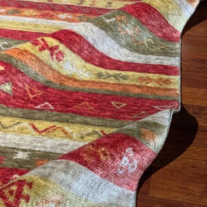 May include: Close-up of a colorful rug with a rolled-up edge. The rug features horizontal stripes in shades of red, yellow, orange, and green. The design includes small geometric and floral patterns. The rug is made of a soft, textured material.