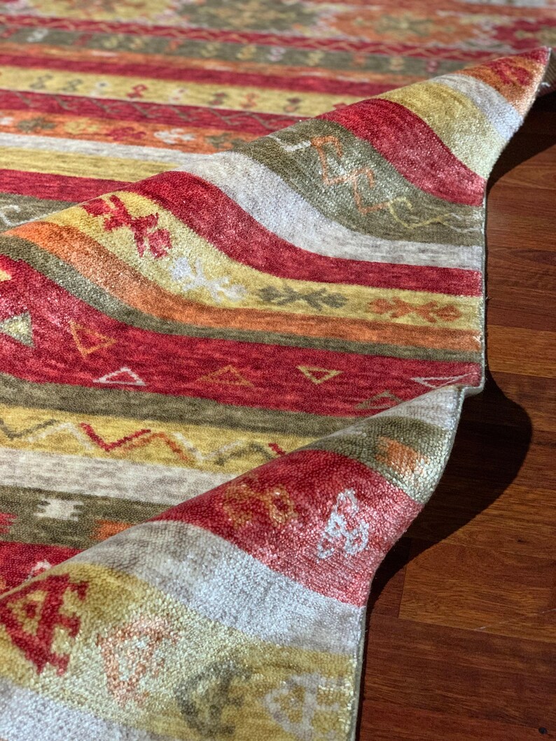 May include: Close-up of a colorful rug with a rolled-up edge. The rug features horizontal stripes in shades of red, yellow, orange, and green. The design includes small geometric and floral patterns. The rug is made of a soft, textured material.