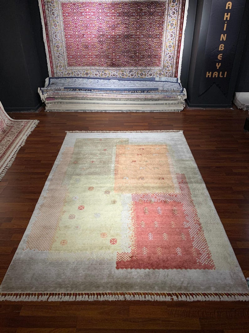 May include: A rectangular area rug with a muted color palette. The rug features a light beige background with sections of peach, orange, and red. The design includes geometric shapes and patterns. The rug has a fringe along the bottom edge.