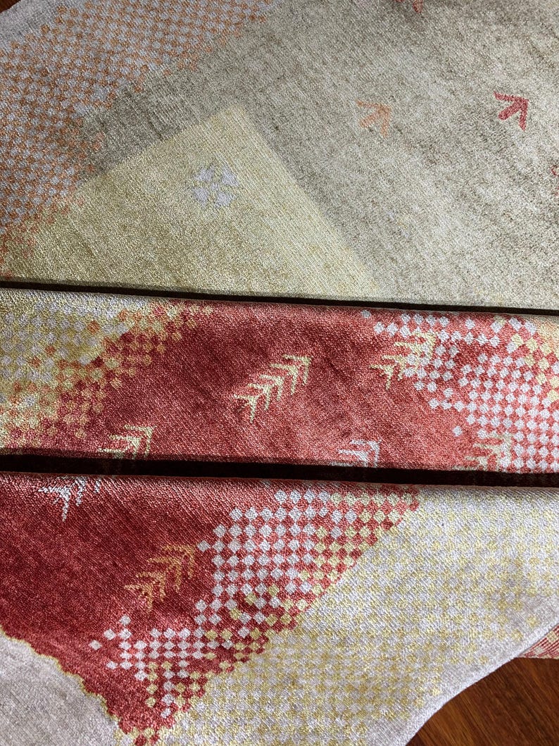 May include: Close-up of a patterned textile with a mix of colors. The design features geometric shapes and arrow-like motifs in shades of red, yellow, and beige. The fabric appears to be folded, revealing layers of the pattern.