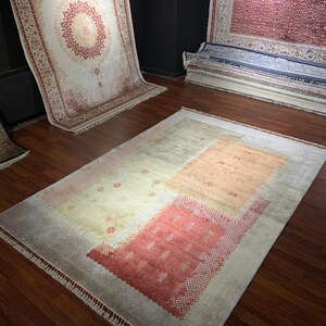 May include: A rectangular rug with a light gray border and a central design featuring peach and red geometric shapes. The rug has a fringe along the edges and is displayed on a hardwood floor. Other rugs are visible in the background.