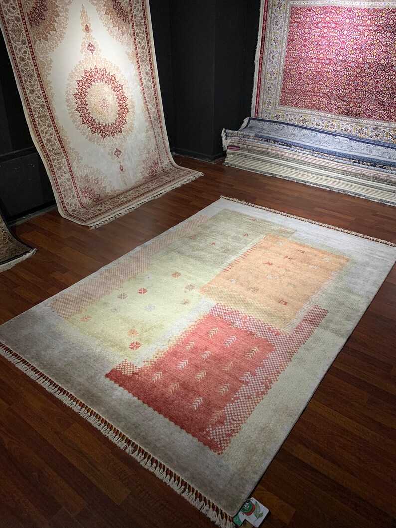 May include: A rectangular rug with a light gray border and a central design featuring peach and red geometric shapes. The rug has a fringe along the edges and is displayed on a hardwood floor. Other rugs are visible in the background.