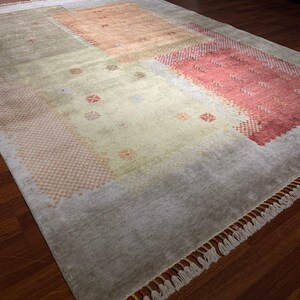 May include: A rectangular area rug with a muted color palette. The rug features a central beige field with peach and red geometric patterns. The border is a light gray with fringe along the edges. The rug is on a dark wood floor.