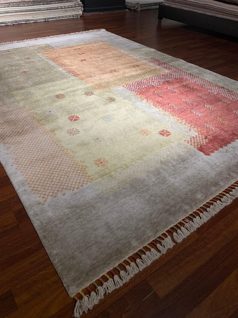 May include: A rectangular area rug with a muted color palette. The rug features a central beige field with peach and red geometric patterns. The border is a light gray with fringe along the edges. The rug is on a dark wood floor.