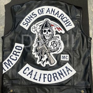 May include: A black leather vest featuring large white embroidered patches with dark blue lettering. The back displays the text SONS OF ANARCHY, CALIFORNIA, and MC, along with a central graphic of a grim reaper holding a rifle and a scythe.