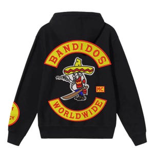 Może przedstawiać: Czarna bluza z kapturem z dużą haftowaną naszywką na plecach. Naszywka przedstawia postać z kreskówki w sombrero trzymającą pistolet i miecz, ze słowami "BANDIDOS WORLDWIDE" w żółto-czerwonym wzorze. Rękaw ma napis "T CY".