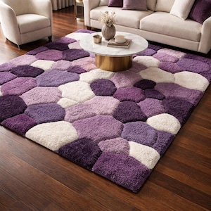 May include: A large, rectangular area rug with a unique pattern of purple, lavender, and white pebble-like shapes. The rug is placed on a dark wood floor, and the room features a sofa, chairs, and a coffee table.