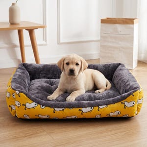 Peut inclure: Un lit pour chien jaune avec un motif de canard et un intérieur en peluche grise. Un chiot Labrador de couleur claire est confortablement allongé dans le lit. Le lit est rectangulaire et a des côtés surélevés.