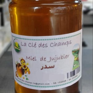 May include: A glass jar of honey with a gold lid. The label reads "La clé des Champs" and "Miel de Jujubier". A cartoon bee is on the label. The honey is a golden amber color.