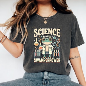 Peut inclure: T-shirt gris foncé avec un graphisme coloré représentant une grenouille en blouse de laboratoire tenant un bécher. Le texte dit "SCIENCE is my SWAMPERPOWER" dans une police ludique, avec des illustrations scientifiques.