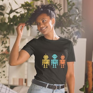 Peut inclure: T-shirt noir avec trois robots colorés : jaune, bleu et orange. Chaque robot porte un numéro : 01, 10 et 11. Le design est simple et graphique, parfait pour un look décontracté.