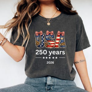 Peut inclure: T-shirt gris foncé avec le mot "USA" dans un design patriotique, avec des éléments rouges, blancs et bleus, des imprimés léopard et des nœuds. Le texte "250 years" et l'année "2026" sont en dessous.