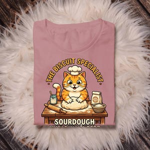 Può includere: T-shirt rosa chiaro con un gatto dei cartoni animati con un cappello da chef, che impasta. Il testo recita "THE BISCUIT SPECIALIST" e "SOURDOUGH". Un mattarello, farina e una ciotola sono sul tavolo di legno.